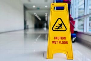 a bright yellow, A-frame Caution Wet Floor sign. The sign has a black triangular symbol depicting a person slipping