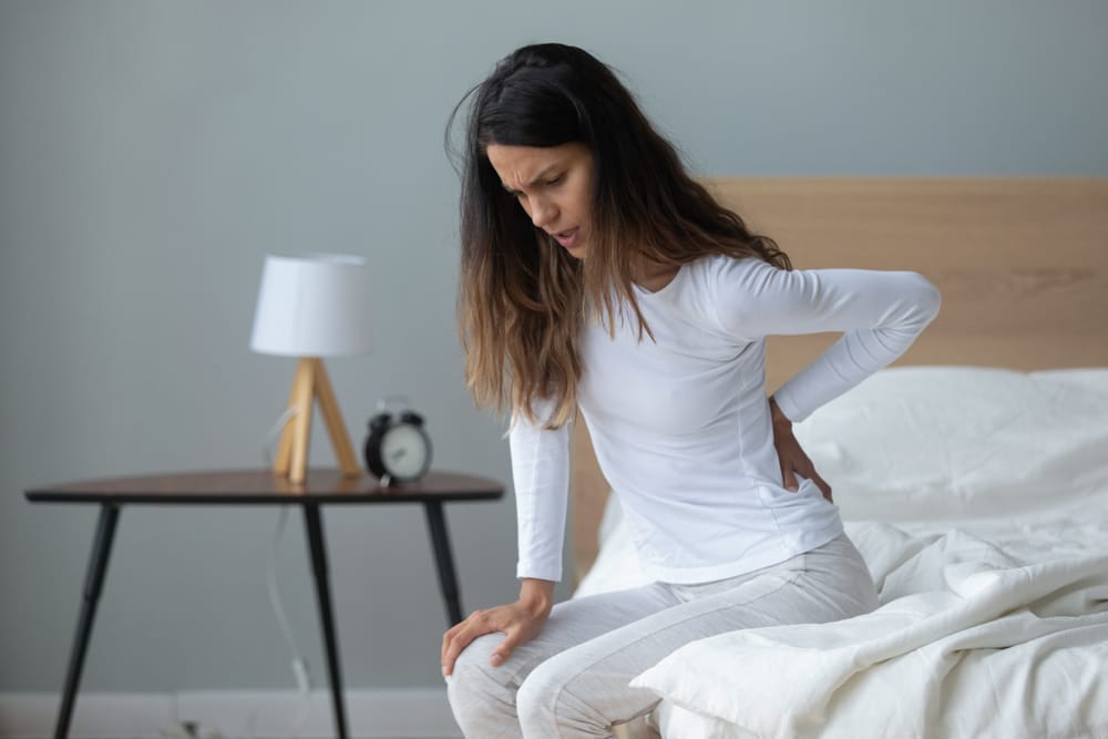 unhappy millennial multiracial woman sitting on bed massaging back suffering from painful feelings waking up in morning after sleeping on uncomfortable mattress at home or in hotel room.